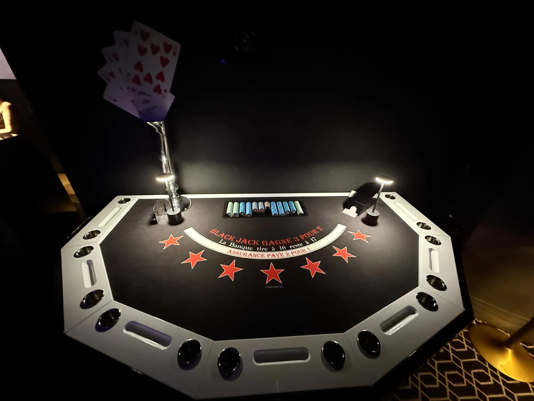 Table de blackjack octogonale blanche sur fond noir, avec une flûte de cartes et les jetons du croupier